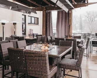 A cozy outdoor patio setup featuring wicker chairs and tables with built-in fire pit features. Snow is gently falling, creating a serene winter atmosphere. The patio is surrounded by protective curtains and heaters are positioned for warmth.