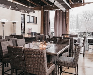 A cozy outdoor patio setup featuring wicker chairs and tables with built-in fire pit features. Snow is gently falling, creating a serene winter atmosphere. The patio is surrounded by protective curtains and heaters are positioned for warmth.
