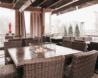 An outdoor seating area with wicker chairs and wooden tables. There are glass-protected fire pits on the tables. The setting is under a partially open roof with curtains, and the background shows snow-covered trees and shrubs. Patio heaters and closed umbrellas are visible, suggesting a cold, possibly winter setting.
