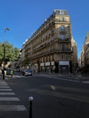 A classic Parisian street scene featuring a traditional Haussmann-style building with ornate balconies and tall windows. The street below has minimal traffic, with a few pedestrians and a car visible. The sky is clear and blue, adding to the peaceful urban ambiance. Green trees line the street, contrasting against the stone facade of the building.
