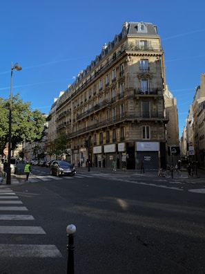 A classic Parisian street scene featuring a traditional Haussmann-style building with ornate balconies and tall windows. The street below has minimal traffic, with a few pedestrians and a car visible. The sky is clear and blue, adding to the peaceful urban ambiance. Green trees line the street, contrasting against the stone facade of the building.