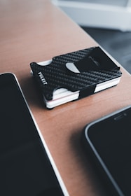 A sleek, black carbon fiber wallet rests on a wooden surface beside two smartphones. The wallet has a minimalist design with visible cards on one side. The setting has a modern and organized feel.