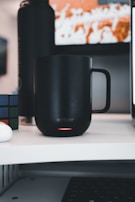 A black smart mug is placed on a white desk, with a subtle red glow at the bottom indicating it's powered on. Next to it, a Rubik's cube is partially visible. In the background, there is a blurred screen with orange and white shapes.