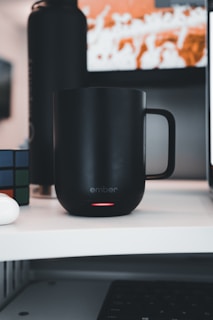 A black smart mug is placed on a white desk, with a subtle red glow at the bottom indicating it's powered on. Next to it, a Rubik's cube is partially visible. In the background, there is a blurred screen with orange and white shapes.