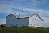 A large white barn with a metal gabled roof sits atop a grassy hill under a partly cloudy blue sky. The barn features multiple ventilation shafts on the top and has a simple, sturdy structure typical of rural farm buildings.