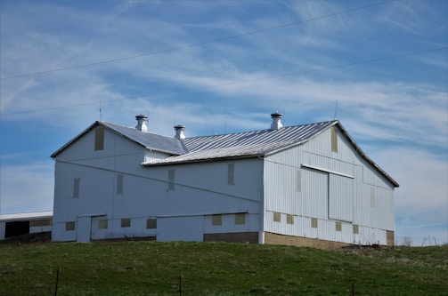 A large white barn with a metal gabled roof sits atop a grassy hill under a partly cloudy blue sky. The barn features multiple ventilation shafts on the top and has a simple, sturdy structure typical of rural farm buildings.
