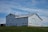 A large white barn with a metal gabled roof sits atop a grassy hill under a partly cloudy blue sky. The barn features multiple ventilation shafts on the top and has a simple, sturdy structure typical of rural farm buildings.