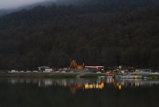 A lakeside scene features a small amusement park with various rides and structures, set against a backdrop of dense, dark forested hills. Several cars are parked along the edge of the amusement area. Bright, colorful lights from the park are reflected in the calm water of the lake, creating a serene yet lively atmosphere.