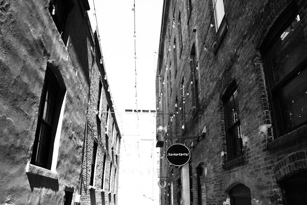 A narrow alleyway lined with brick buildings on both sides. There are hanging string lights strung across the alley, adding a decorative element. A sign with text hangs from one of the buildings. The image is in black and white, highlighting the textures of the brickwork and the light contrasts.