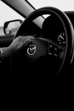A black and white image of a hand gripping the steering wheel of a car, featuring the Mazda logo prominently in the center. The dashboard controls are visible on the right side of the wheel. The driver's side mirror is slightly visible through the window, giving a sense of being inside a parked or moving vehicle.
