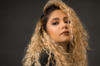 A person with long, curly blonde hair looks confidently towards the camera with a slight smile. The lighting is soft, highlighting their facial features and the texture of their hair. The background is a neutral dark tone, bringing focus to the subject.