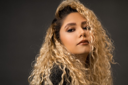 A person with long, curly blonde hair looks confidently towards the camera with a slight smile. The lighting is soft, highlighting their facial features and the texture of their hair. The background is a neutral dark tone, bringing focus to the subject.