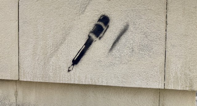 Stenciled graffiti of a microphone is spray-painted on a textured concrete wall. The graffiti is executed in black, standing out against the light gray background of the concrete surface.