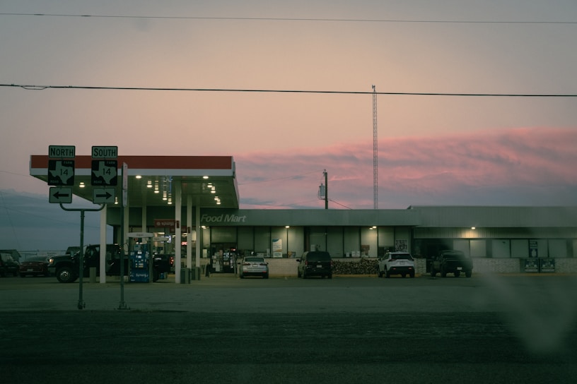 A gas station and convenience store, labeled 'Food Mart,' is situated at a crossroads. Several vehicles are parked near the pumps, and directional road signs indicate routes North and South for Highway 14. The sky displays a soft pink hue during what appears to be sunset or sunrise.