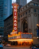 A brightly lit marquee sign of the Chicago Theatre features prominently against a backdrop of urban buildings. The sign displays the name 'Chicago' vertically in bright red and white lights. Below the main sign, a marquee board announces upcoming events, including performances by the Tedeschi Trucks Band and others. Pedestrians and cars pass by on the street in front of the historic theatre building, which exhibits intricate architectural details.