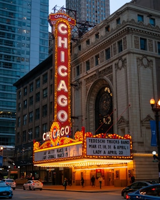 A brightly lit marquee sign of the Chicago Theatre features prominently against a backdrop of urban buildings. The sign displays the name 'Chicago' vertically in bright red and white lights. Below the main sign, a marquee board announces upcoming events, including performances by the Tedeschi Trucks Band and others. Pedestrians and cars pass by on the street in front of the historic theatre building, which exhibits intricate architectural details.