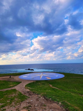 A circular, compass-like design with various symbols is situated on a grassy area. It is positioned near the edge of a cliff overlooking the ocean. The sky is partially cloudy with various shades of blue and white clouds, and the sunlight filters through creating a serene, picturesque view.