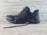 A black athletic shoe with a rugged sole and mesh detailing, designed for outdoor activities. It features brand markings on the side and is positioned against a light wooden background.