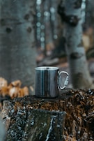 A metal mug sits on a tree stump in a forest setting. The background is blurred, showing tall trees with a cool color palette. Snowflakes appear to be falling softly around the scene, creating a serene and quiet atmosphere.