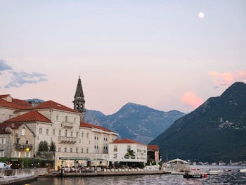 A picturesque coastal town with a large building featuring red-tiled roofs and a stone tower set against a backdrop of mountains during sunset. The waterfront has small boats and a reflective, calm body of water.