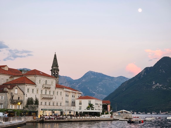 A picturesque coastal town with a large building featuring red-tiled roofs and a stone tower set against a backdrop of mountains during sunset. The waterfront has small boats and a reflective, calm body of water.