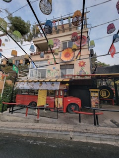 A multi-story building with vibrant murals on its facade, including large flowers and a bicycle. Colorful lanterns and baskets hang from overhead wires, adding a festive atmosphere. In front of the building, a red bus modified into a cafe or food truck is parked, sheltered by a glass structure. The area is surrounded by greenery and has a casual, artistic vibe.