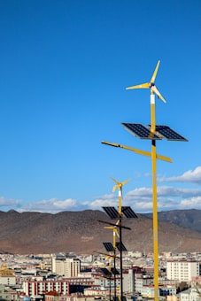 A line of yellow wind turbines with attached solar panels is situated in front of a cityscape. The background features dry, mountainous terrain under a clear blue sky with a few scattered clouds.