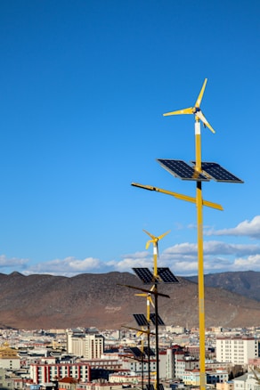 A line of yellow wind turbines with attached solar panels is situated in front of a cityscape. The background features dry, mountainous terrain under a clear blue sky with a few scattered clouds.