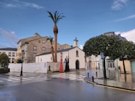 A small chapel is seen at a street corner, surrounded by traditional European buildings under a partly cloudy sky. Two statues depicting figures, one wearing a red dress and another in a black suit, are positioned beside a tall palm tree. The wet pavement suggests recent rainfall, and a pedestrian crossing sign can be observed near a tree.