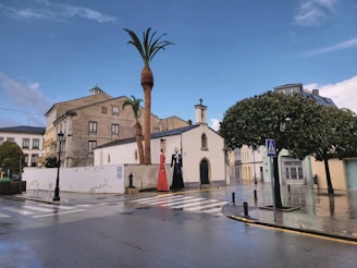 A small chapel is seen at a street corner, surrounded by traditional European buildings under a partly cloudy sky. Two statues depicting figures, one wearing a red dress and another in a black suit, are positioned beside a tall palm tree. The wet pavement suggests recent rainfall, and a pedestrian crossing sign can be observed near a tree.