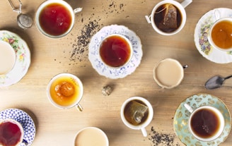 Multiple cups of tea are arranged on a wooden surface, each in a different and uniquely decorated teacup. There are various types of teas, some with tea bags, some with milk, and some plain. Loose tea leaves are scattered around the cups.