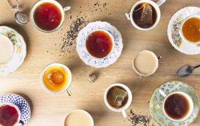 Multiple cups of tea are arranged on a wooden surface, each in a different and uniquely decorated teacup. There are various types of teas, some with tea bags, some with milk, and some plain. Loose tea leaves are scattered around the cups.