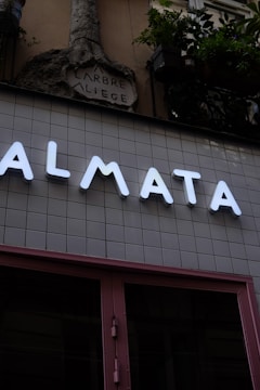 A modern storefront with large illuminated letters spelling out 'ALMATA' against a tiled wall. Above this, an ornate stone sculpture with the words 'ARBRE A LIEGE' is visible. Potted plants hang from the upper part of the structure, adding a touch of greenery.