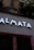 A modern storefront with large illuminated letters spelling out 'ALMATA' against a tiled wall. Above this, an ornate stone sculpture with the words 'ARBRE A LIEGE' is visible. Potted plants hang from the upper part of the structure, adding a touch of greenery.