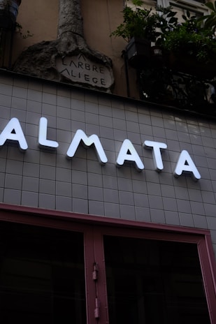 A modern storefront with large illuminated letters spelling out 'ALMATA' against a tiled wall. Above this, an ornate stone sculpture with the words 'ARBRE A LIEGE' is visible. Potted plants hang from the upper part of the structure, adding a touch of greenery.