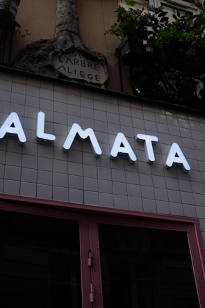 A modern storefront with large illuminated letters spelling out 'ALMATA' against a tiled wall. Above this, an ornate stone sculpture with the words 'ARBRE A LIEGE' is visible. Potted plants hang from the upper part of the structure, adding a touch of greenery.