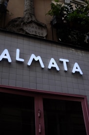 A modern storefront with large illuminated letters spelling out 'ALMATA' against a tiled wall. Above this, an ornate stone sculpture with the words 'ARBRE A LIEGE' is visible. Potted plants hang from the upper part of the structure, adding a touch of greenery.