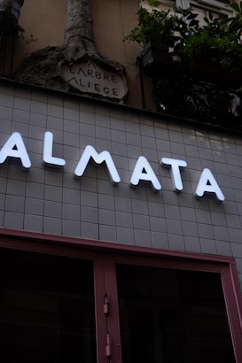 A modern storefront with large illuminated letters spelling out 'ALMATA' against a tiled wall. Above this, an ornate stone sculpture with the words 'ARBRE A LIEGE' is visible. Potted plants hang from the upper part of the structure, adding a touch of greenery.