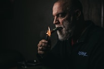 A man with a grey beard is lighting a cigar with a lighter. The scene is dimly lit, creating a moody atmosphere. He is wearing a dark jacket with an inscription on it, and is gazing intently at what he is doing.