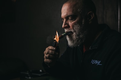 A man with a grey beard is lighting a cigar with a lighter. The scene is dimly lit, creating a moody atmosphere. He is wearing a dark jacket with an inscription on it, and is gazing intently at what he is doing.