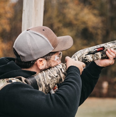 a man with a hat and glasses aims a shotgun