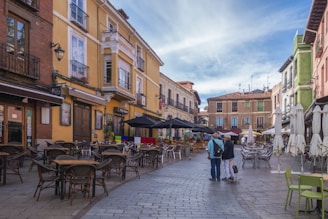 A European street scene features colorful buildings with balconies, a cobblestone pathway lined with outdoor café tables and chairs, and two people walking together in the middle of the walkway. Umbrellas cover some of the seating areas, and the sky above is partly cloudy.