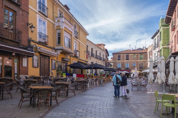 A European street scene features colorful buildings with balconies, a cobblestone pathway lined with outdoor café tables and chairs, and two people walking together in the middle of the walkway. Umbrellas cover some of the seating areas, and the sky above is partly cloudy.