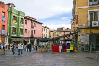 A vibrant urban square with colorful buildings and an outdoor seating area. People are walking around and engaging in conversation. There are several potted plants and a stand with a geometric design. The sky is partly cloudy.
