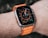 A close-up view of a smart watch with a round, digital display showing various metrics and a colorful interface. The watch is secured with an orange strap and is worn on a wrist with visible water droplets, suggesting recent exposure to moisture.