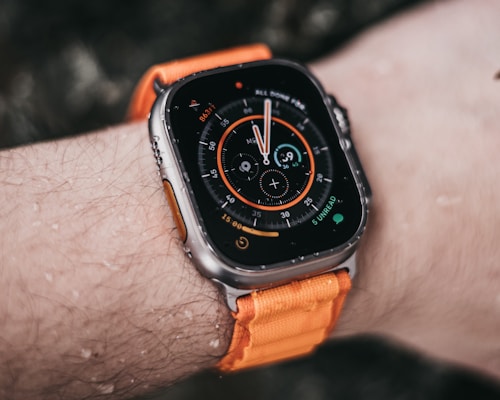 A close-up view of a smart watch with a round, digital display showing various metrics and a colorful interface. The watch is secured with an orange strap and is worn on a wrist with visible water droplets, suggesting recent exposure to moisture.