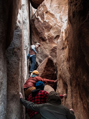 Four individuals are climbing through a narrow rocky crevice, with one person leading the way at a slightly higher altitude. The rocks are large and imposing, creating a sense of adventure and challenge.