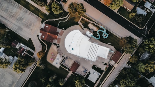 An aerial view of a recreational area featuring a large, rectangular swimming pool with marked lanes and a small water slide. Surrounding the pool are pathways, green lawns, and adjacent buildings with brown roofs. Trees and parking lots border the area, creating a contrast between natural and urban elements.