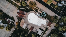 An aerial view of a recreational area featuring a large, rectangular swimming pool with marked lanes and a small water slide. Surrounding the pool are pathways, green lawns, and adjacent buildings with brown roofs. Trees and parking lots border the area, creating a contrast between natural and urban elements.
