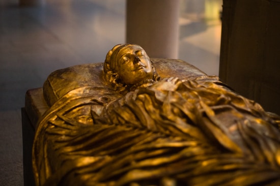 A detailed sculpture of a reclining figure, crafted from gold or a gold-like material. The figure appears serene, with closed eyes and hands resting on the chest. The intricate folds of the draped garment and the serene facial expression convey a sense of peacefulness. Soft lighting emphasizes the reflective surface of the sculpture.